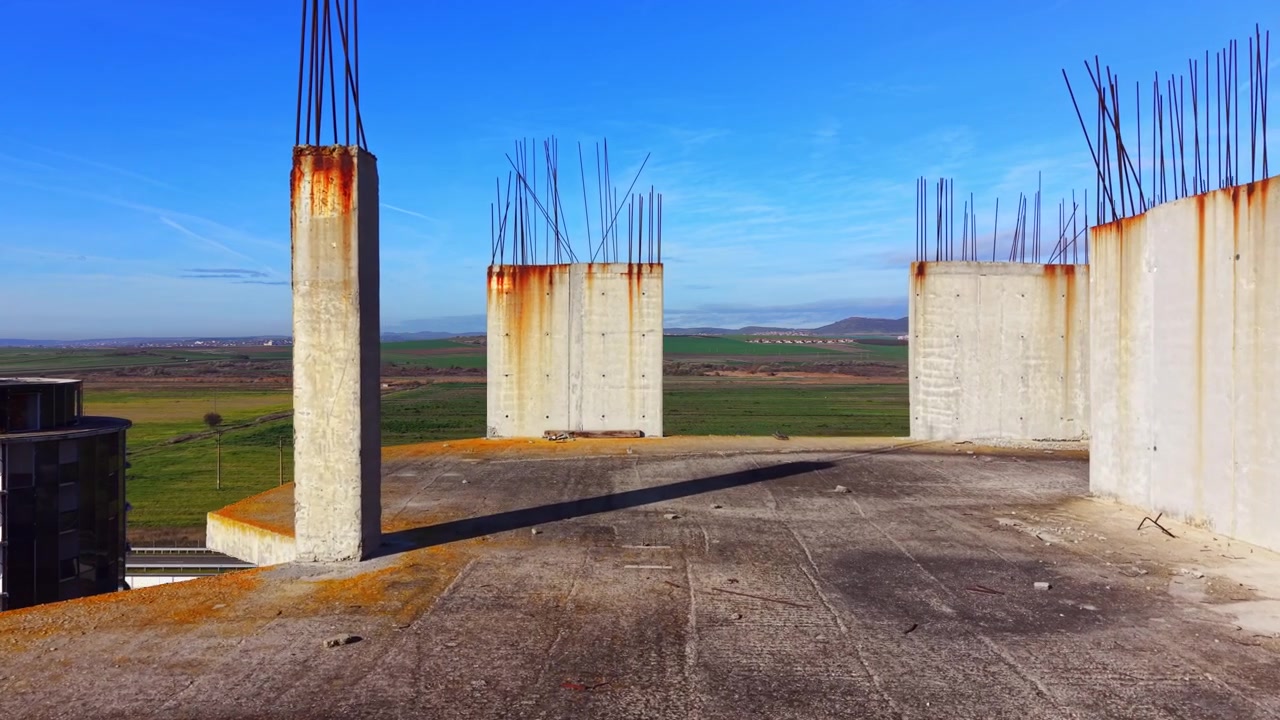 File di colonne di cemento si ergono sopra un cantiere costiero. La prospettiva dall'alto rivela punte di tondini arrugginiti, una torre di vetro e terreni agricoli verdi oltre.
