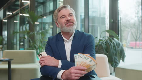 successful businessman holding smartphone and counting euro bill money fan sitting in office lobby