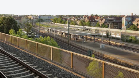 vías ferroviarias en primer plano que conducen hacia una estación de tren en la hora dorada, con pasajeros esperando y un paisaje urbano de fondo