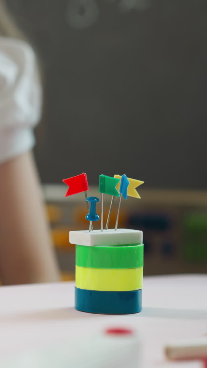 Little girl builds tower with eraser and small flags on top at desk in classroom closeup. Schoolgirl has fun with color stationery at craft lesson