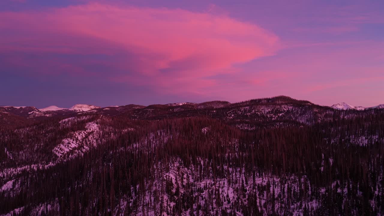 Ripresa aerea con formazioni nuvolose spettacolari che risplendono di rosa e viola sulle innevate San Juan Mountains. Il cielo vivace contrasta con la cresta della pineta scura, creando una surreale atmosfera invernale.