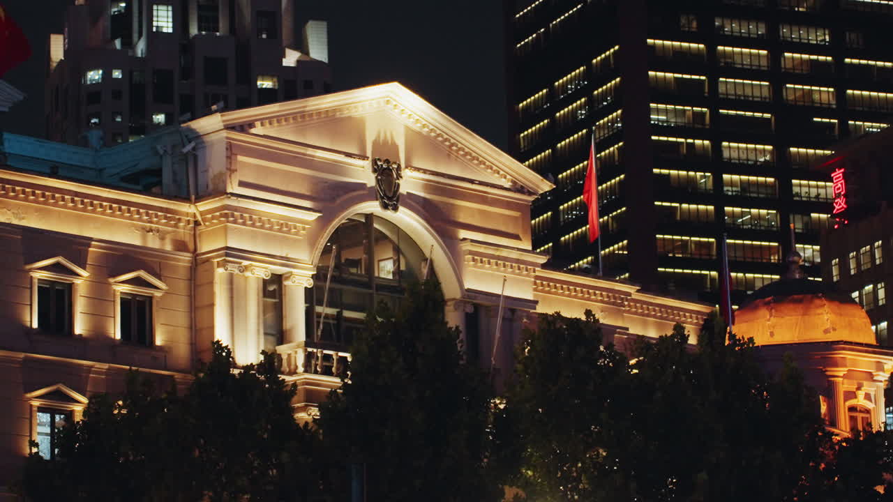 Facciata neoclassica dorata con arco e colonne, ingresso ad arco immerso in una luce calda. Una torre di vetro adiacente si erge sullo sfondo, mentre la silhouette di alberi e bandiere incornicia la scena, creando un'atmosfera serale vivace.