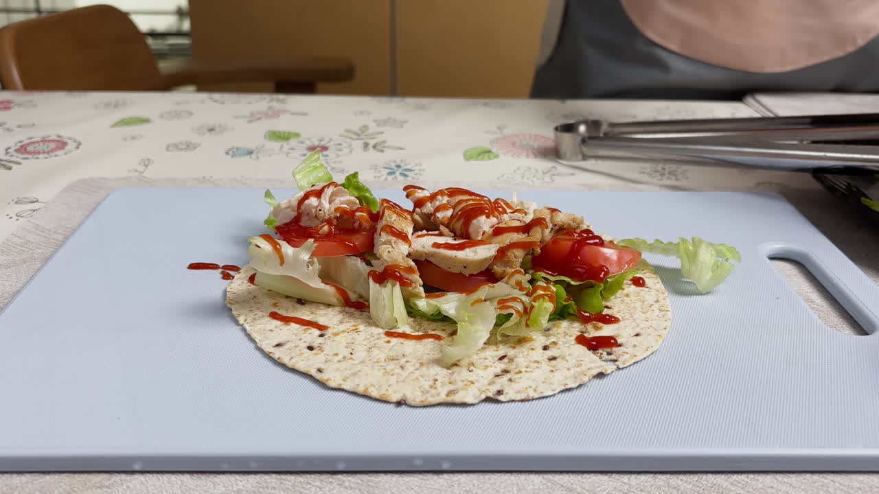 Fresh chicken wrap on tortilla with vegetables being topped with ketchup and mayonnaise, homemade meal preparation in kitchen