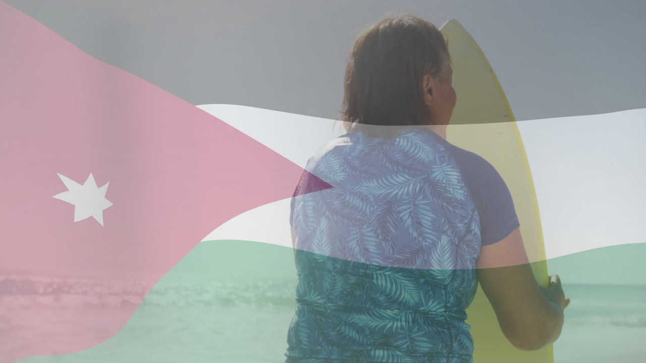 Holding surfboard with Jordanian flag design, person standing by ocean waves