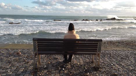 una persona se sienta sola en un banco junto al mar viendo las olas furiosas romper en tierra