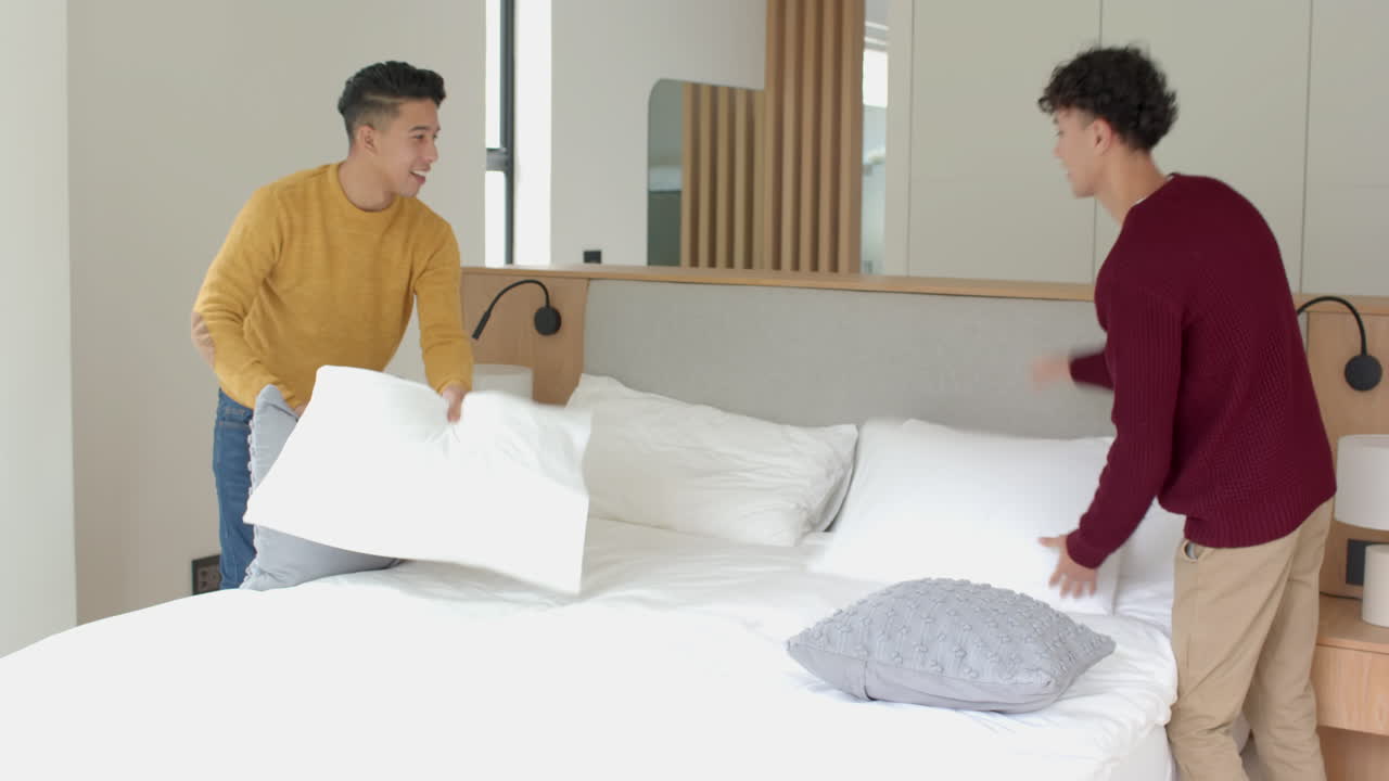 Making bed , multiracial gay couple arranging pillows in modern bedroom, at home