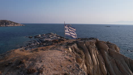 national flag of greece waving on pole on top of cliff above aegean sea, drone shot