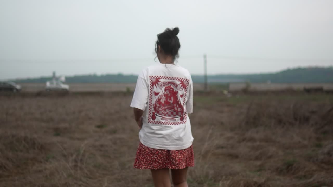 Giovane donna che cammina attraverso un campo secco, con una camicia bianca e pantaloncini rossi, vista di spalle.