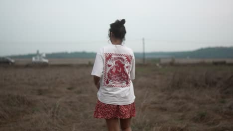 Young-woman-walking-through-a-dry-field-wearing-a-white-shirt-and-red-shorts,-rear-view