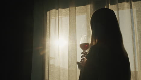 a woman with a glass of red wine stands at the window at sunset