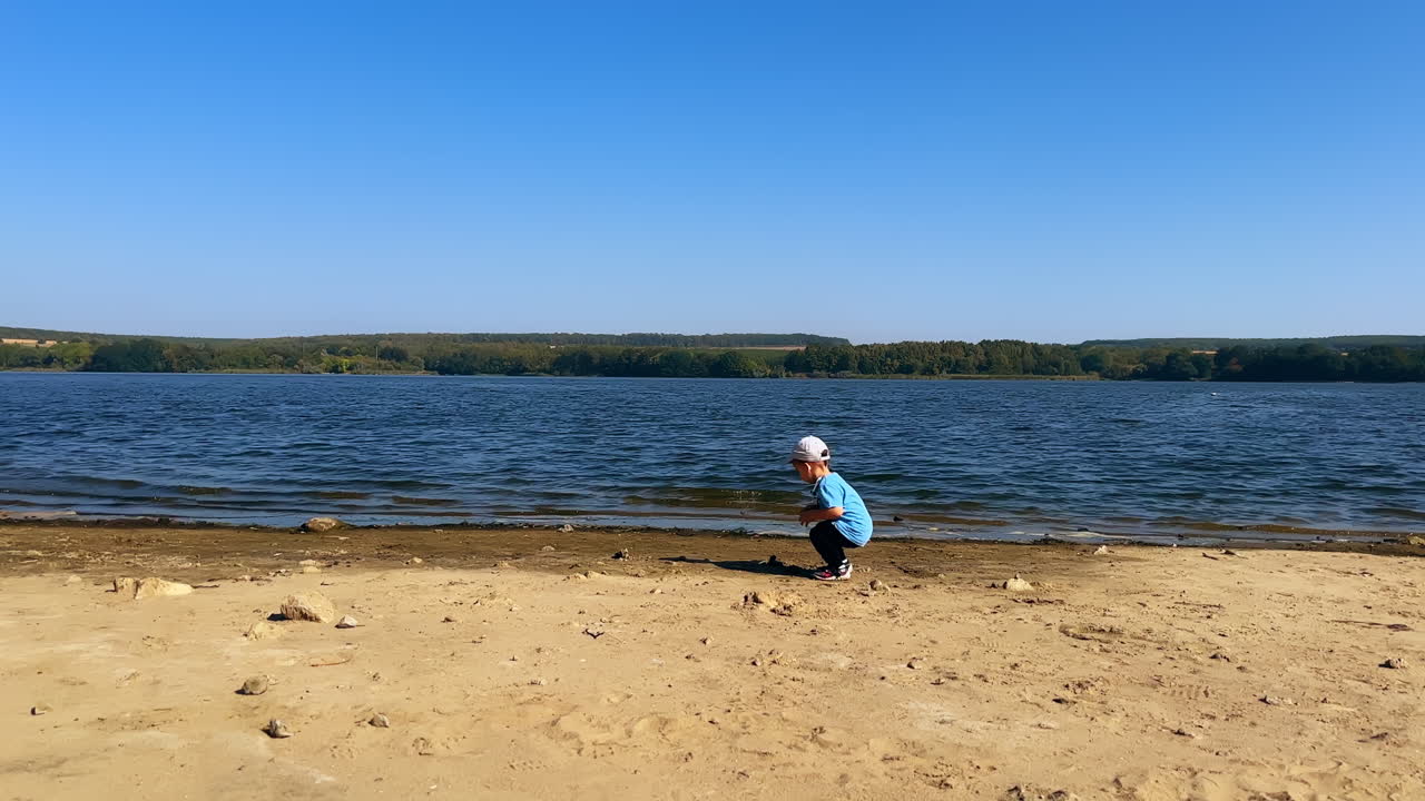 Little kid in a cap walks by the river bank. Baby boy takes the stone and throws it into water.