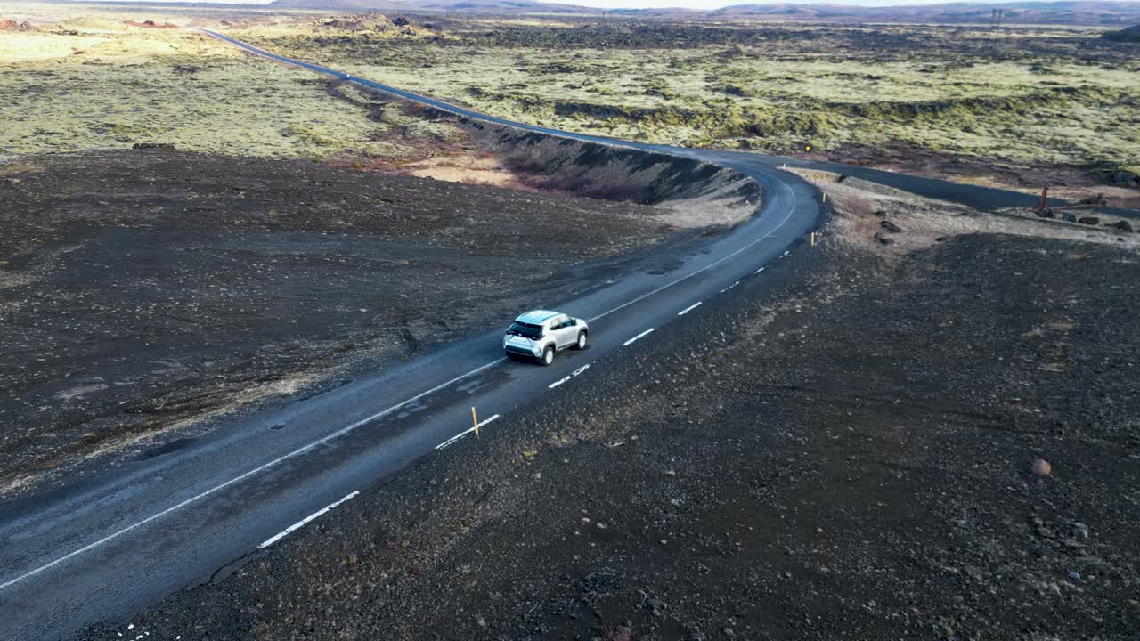 um carro em uma estrada sinuosa nas terras altas da islândia, passando por campos de musgo e colinas, chegando a uma vasta planície