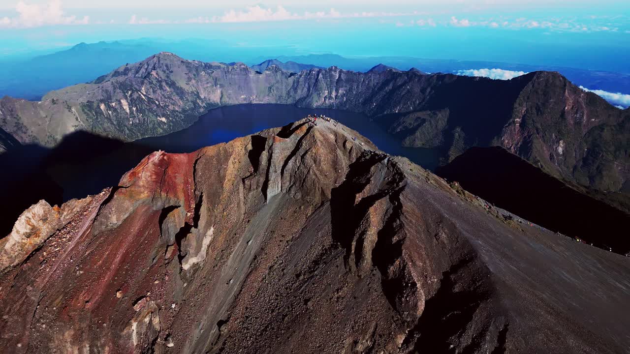 Aerial drone shot of trekkers on Mount Rinjani peak surrounded by volcanic landscape and sparkling crater lake under beautiful natural light.