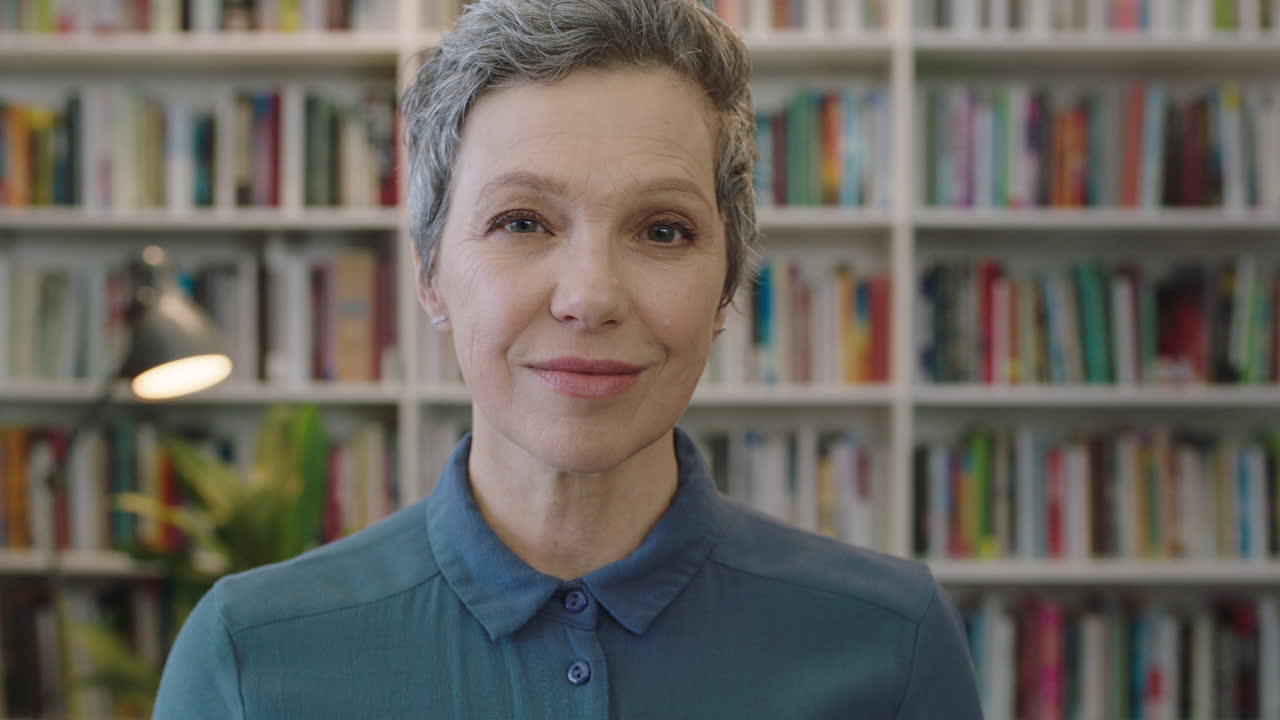 ritratto di una donna caucasica matura e amichevole bibliotecaria sorridente che guarda con fiducia alla telecamera sullo sfondo della biblioteca