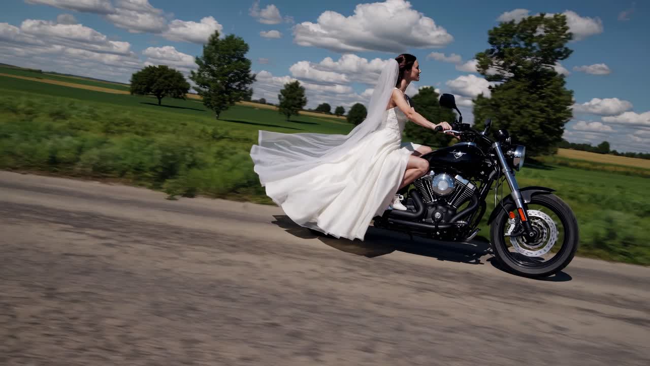 Radiant bride wearing flowing white wedding gown riding sleek black motorcycle along scenic countryside road, green fields and cloudy blue sky creating dramatic backdrop of freedom and adventure