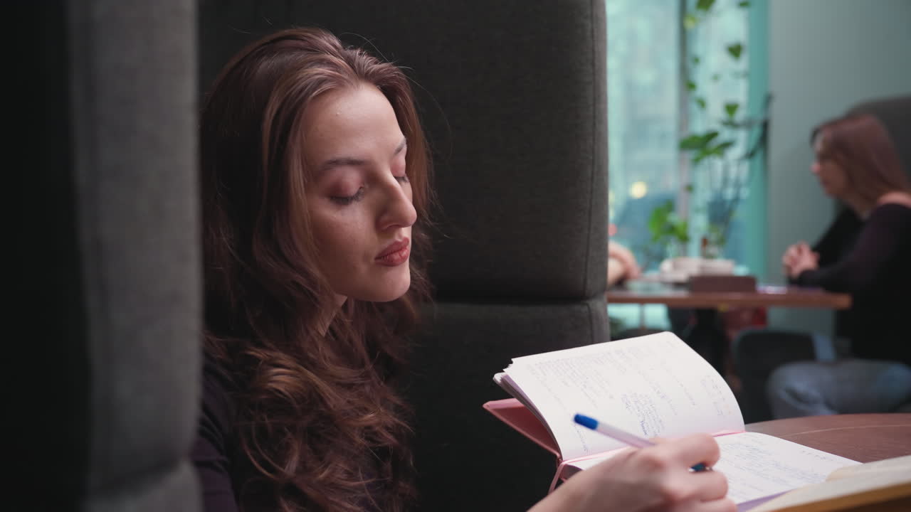 Side view of young woman with long brown hair writing in notebook with focused expression while seated in booth, blurred background shows people at table and greenery outside window, indoor study vibe