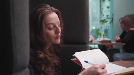 side view of young woman with long brown hair writing in notebook with focused expression while seated in booth, blurred background shows people at table and greenery outside window, indoor study vibe