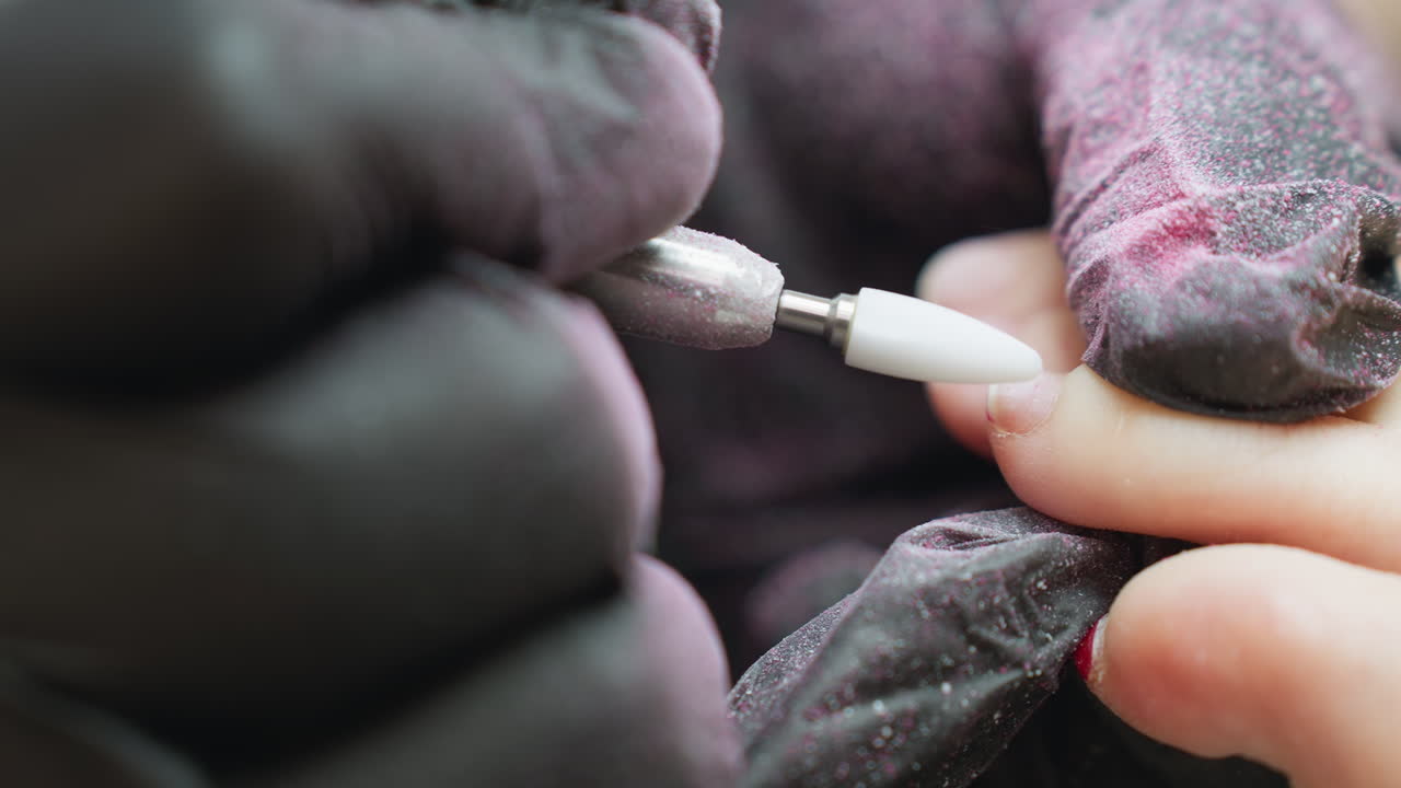 Close up of nail technician in black glove using electric file on client toe nail while red polish particles cover glove and float in air as technician transitions to next toe