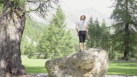 Little-girl-climbs-on-a-rock-and-spreads-her-arms-towards-the-mountain,-forest-of-Italian-Alps-4K