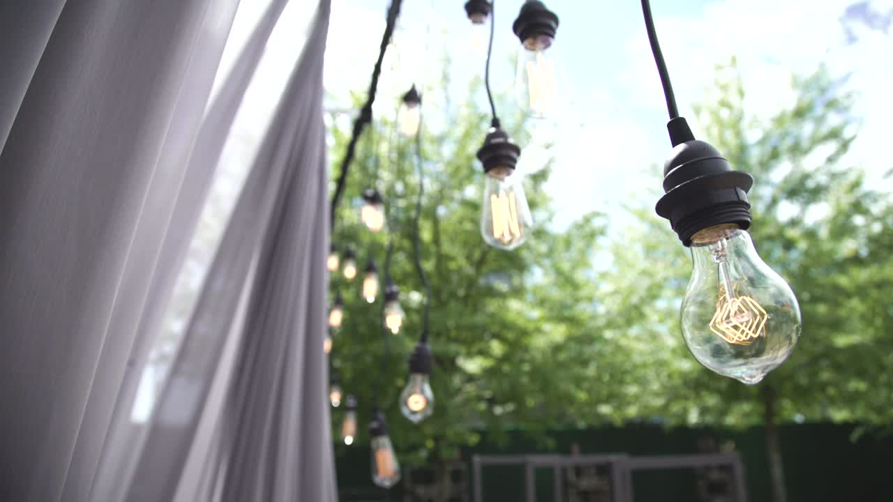 4k 비디오 전구 에디슨 유선 빈티지 스타일은 자연에서 이벤트 중에 매달려 있습니다. 초점과 초점 밖에서 느린 동작으로 촬영 된 영화적 스타일의 웨딩 이벤트 의식, 야외 이벤트.
