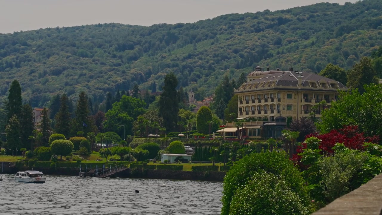 Primo piano di una storica villa di lusso sul Lago Maggiore con una barca che riposa sull'acqua calma e auto che passano lungo la strada lungolago, catturando l'autentico fascino lacustre italiano