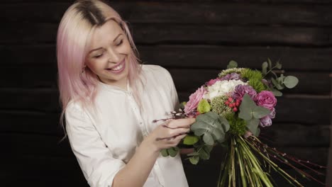 a pink haired young woman makes the design of the bouquet, complementing the composition with willow branches. smiling. dark background