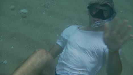 adulto joven buceando con esnórquel y gafas sobre una playa blanca bajo el agua y saludando