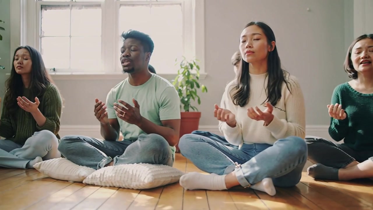 Panorerande kamera visar sittande vuxna som sjunger för meditation i ett ljust övningsrum, kuddar