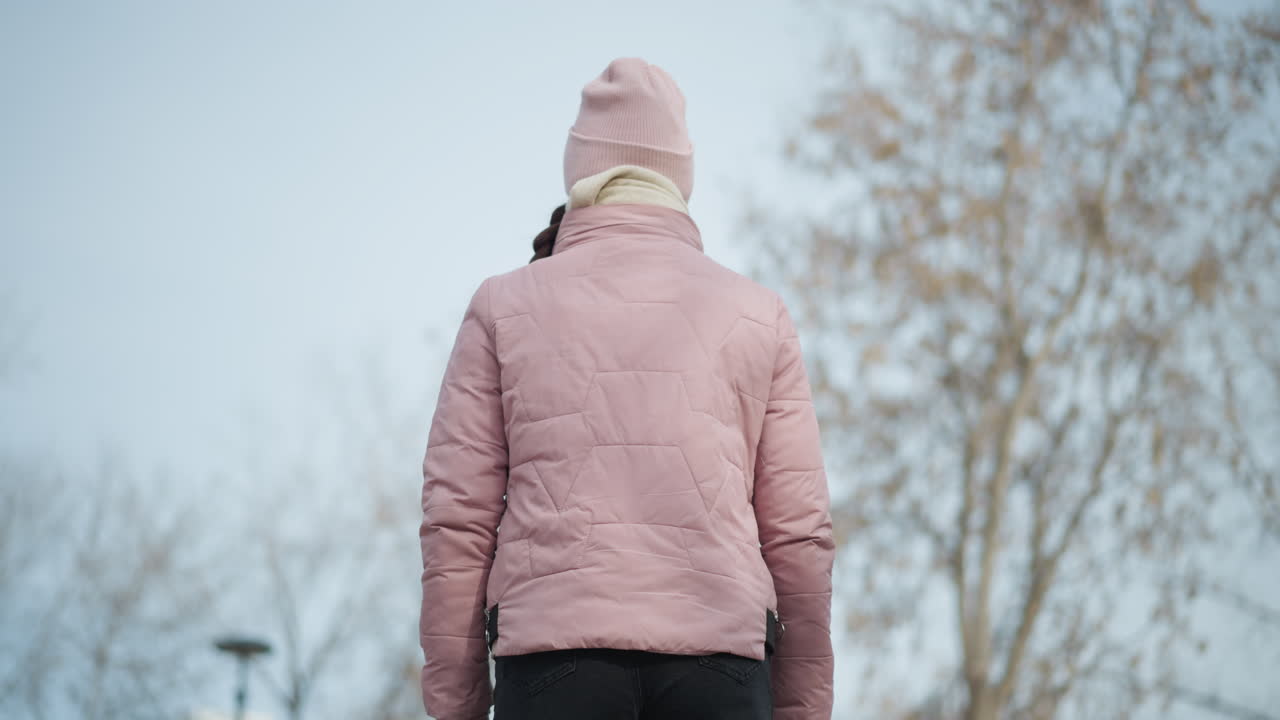 Woman in pink winter jacket and knit hat seen from behind, walking outdoors on chilly day beneath soft blue sky, surrounded by bare trees, embodying solitude, calm, seasonal change and quiet reflection