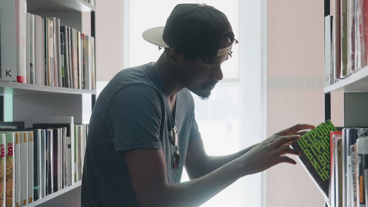 Side view young man wearing cap and casual shirt browsing bookshelf in bright modern library, carefully retrieving book, examining cover, and returning it to shelf with focused