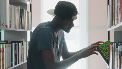 side view young man wearing cap and casual shirt browsing bookshelf in bright modern library, carefully retrieving book, examining cover, and returning it to shelf with focused