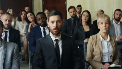 front view of caucasian businessman sitting among people in a conference room raising his hand talking to the camera