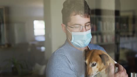 caucasian man wearing face mask, standing at window and petting dog