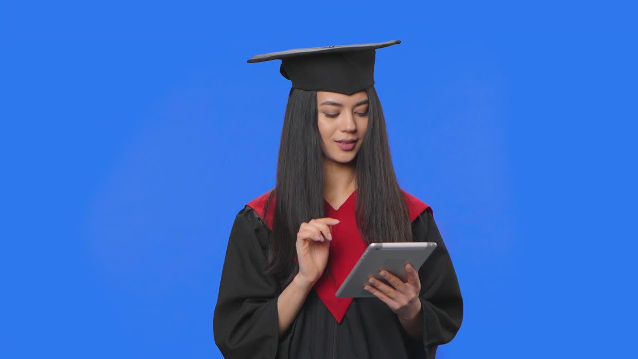retrato de una estudiante en traje de graduación de gorra y túnica, viendo información en una tableta digital. mujer joven posando en el estudio con fondo de pantalla azul. primer plano. cámara lenta lista 59.94fps