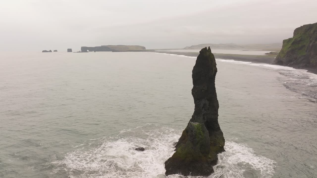 A lone Reynisdrangar sea stack rises from the ocean off Reynisfjara beach in southern Iceland, with black sand beaches and rugged cliffs in the background.