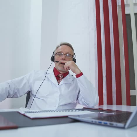 physician with headset talks in medical office. professional male doctor with headphones in white medical uniform sitting at desk and speaking into microphone in modern hospital office.