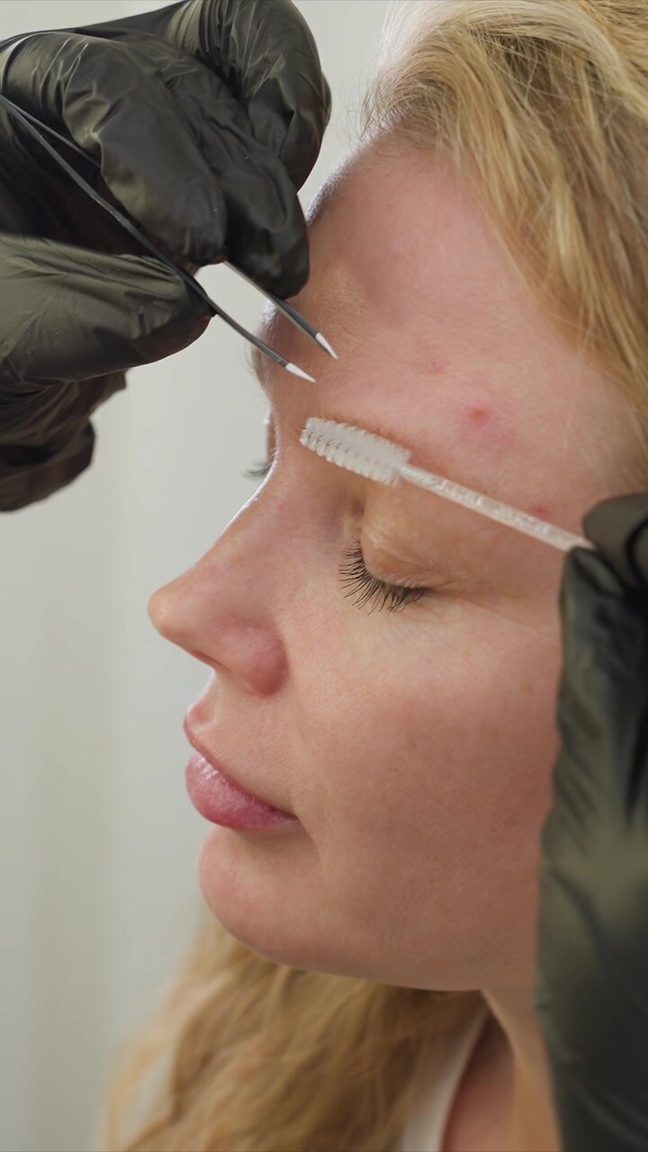 Close up sequence shows beauty professional performing eyebrow grooming service on client. focus remains tightly on detailed process of tweezing and applying brush with steady interaction