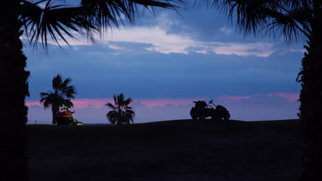 mini tractors with headlights cutting grass at golf coarse in early blue hour as palm tree silhouettes waving in gentle wind