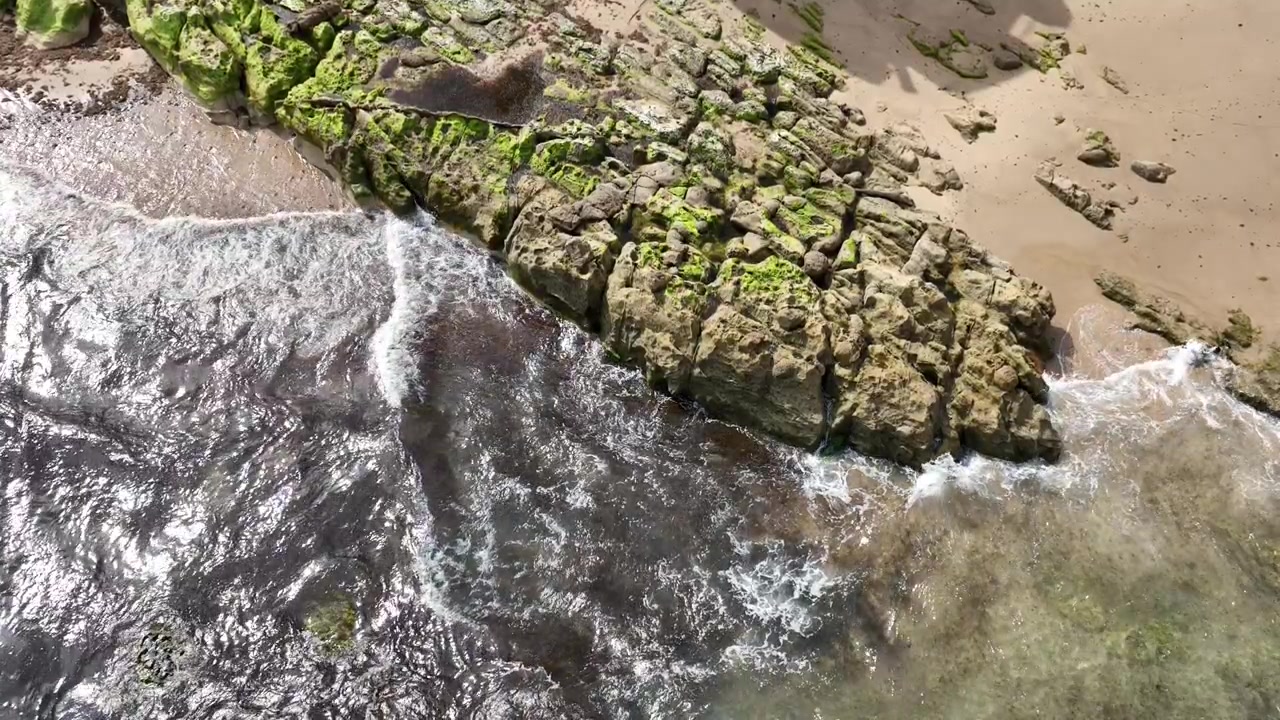 Vue aérienne au ralenti des vagues de l'Atlantique déferlant sur des rochers côtiers accidentés recouverts d'algues vertes près d'Asilah, au Maroc, capturant le mouvement détaillé de l'océan et les textures naturelles du littoral.