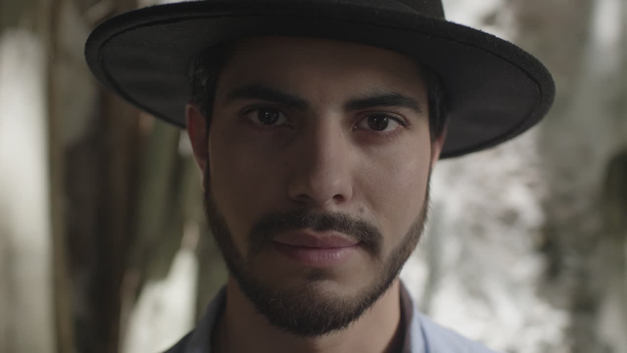 High-quality 4K footage of a stylish young man with a beard and hat, slowly opening his eyes in soft, natural light. He has a focused, mysterious expression. Ideal for travel, discovery