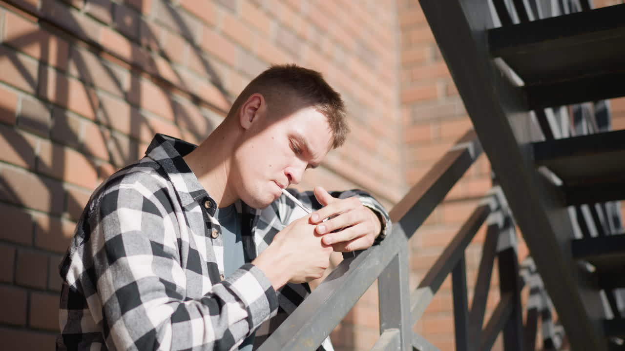 young student resting on iron railing holds cigarette in mouth while igniting flame from lighter held near cigarette pack wearing checkered shirt and jeans under sunlit brick wall and metal staircase