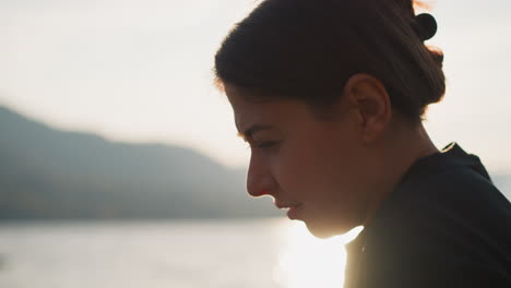 woman sitting on bank at sunset. upset female thinking about sad events in life with view of lake and mountains in sunny weather. pain of being alone surrounded by natural beauty