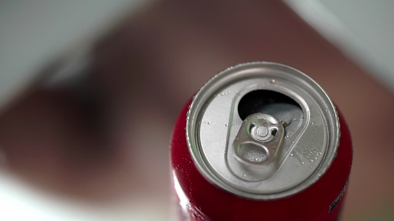 Extreme close-up of cold red soda can with droplets as hand picks it up from reflective surface