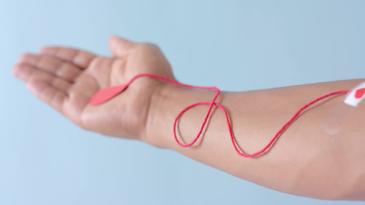 Mano di uomo birazziale con goccia di sangue, filo rosso e cerotto a forma di cuore su sfondo blu, al rallentatore.