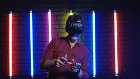 man in vr glasses and playing videogame with joystick in a room with colorful neon lamps on the wall