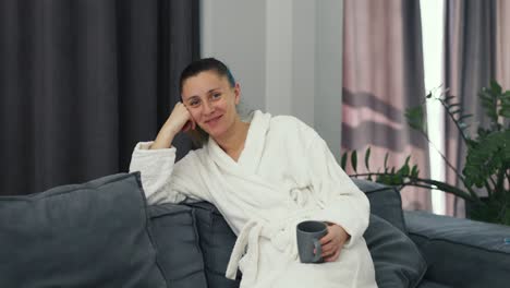 young woman wearing white bathrobe sitting on sofa with cup of tea