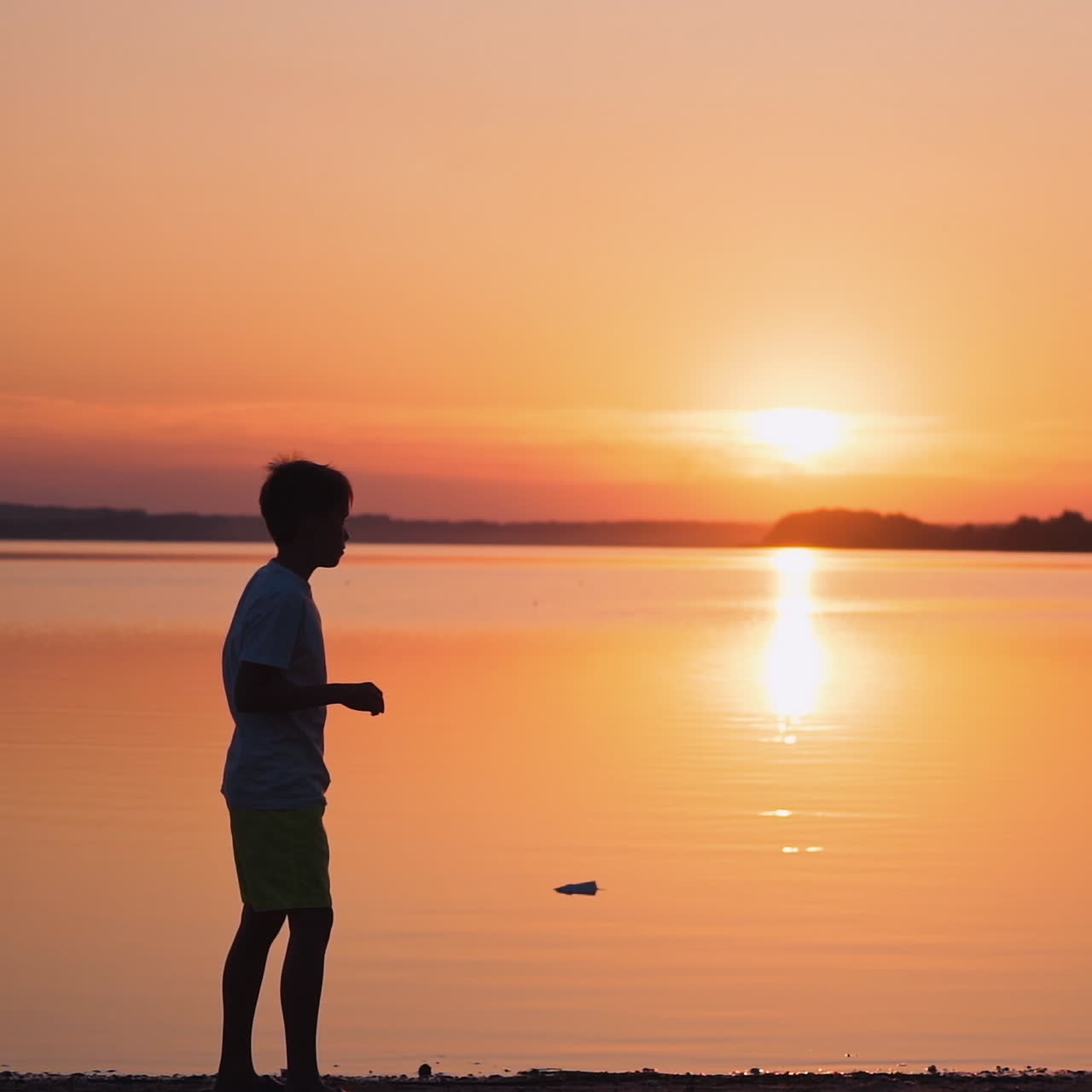Boy near the river at sunset. Side view of a kid who is throwing paper plane up on the beautiful orange background of calm lake. Slow motion.