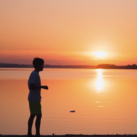 boy near the river at sunset. side view of a kid who is throwing paper plane up on the beautiful orange background of calm lake. slow motion.