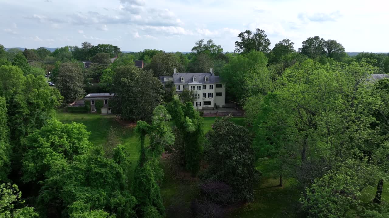 Aerial approaching shot of luxury mansion in green forest landscape of Virginia, United States. Large green garden with grass in suburb. Sunny day with clouds at sky in May.