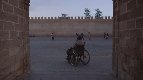 Wheelchair-bound-man-watches-young-men-play-soccer-football-in-public-square-nearby-medina-in-Essaouira-Morocco,-handheld-shot